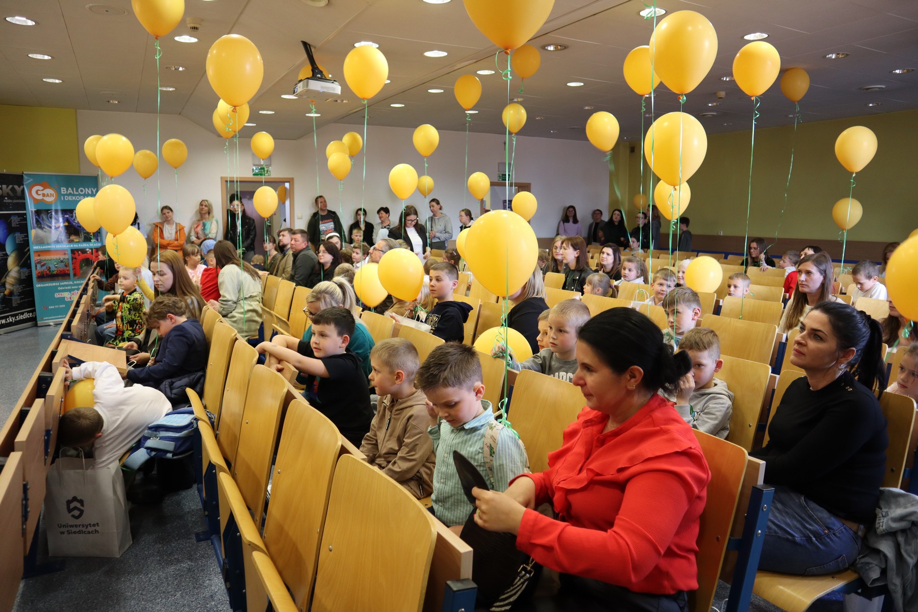 Uczestnicy konkursu na auli z żółtymi balonami unoszącymi się nad nimi